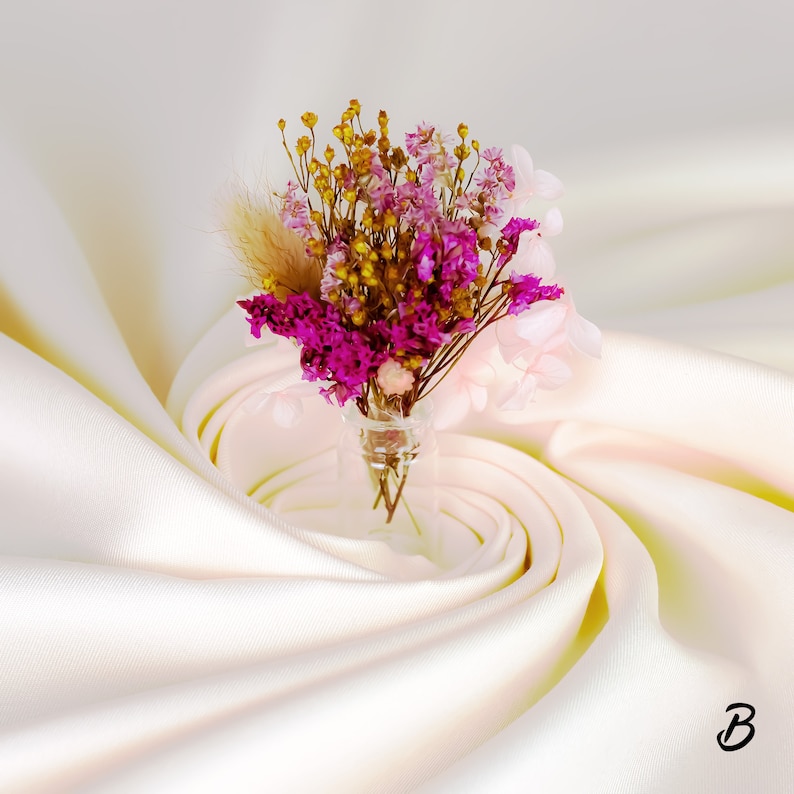 May include: A small glass vase filled with a bouquet of dried flowers. The arrangement includes vibrant magenta, yellow, and pale pink blooms, with a touch of beige. The vase sits on a cream-colored, draped fabric.