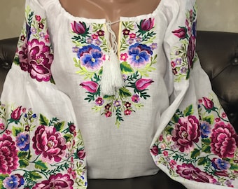 May include: White linen blouse with embroidered floral design in shades of pink, purple, blue, and green. The embroidery features a large floral motif on the front and smaller floral motifs on the sleeves.