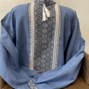 May include: A blue long-sleeved shirt with detailed embroidery in white, black, and blue. The shirt has a high collar and a decorative tassel. The sleeves are long, and the shirt is made of a soft fabric. The shirt is displayed on a chair.
