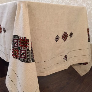 May include: A beige linen tablecloth with embroidered geometric patterns in red, black, and green. The tablecloth has a subtle sheen and a slightly textured surface.