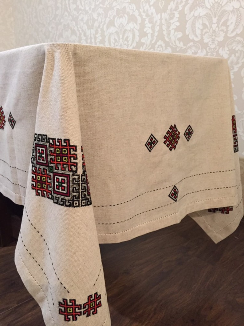 May include: A beige linen tablecloth with embroidered geometric patterns in red, black, and green. The tablecloth has a subtle sheen and a slightly textured surface.