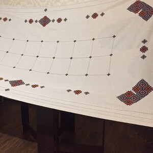 May include: White tablecloth with a geometric embroidery design in red, black, and blue. The design features a grid pattern with repeating squares and diamonds.