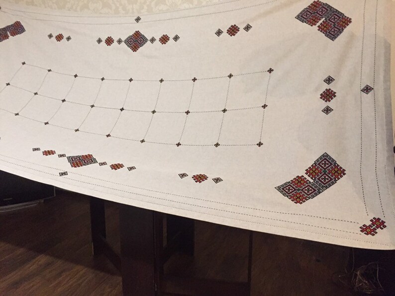 May include: White tablecloth with a geometric embroidery design in red, black, and blue. The design features a grid pattern with repeating squares and diamonds.