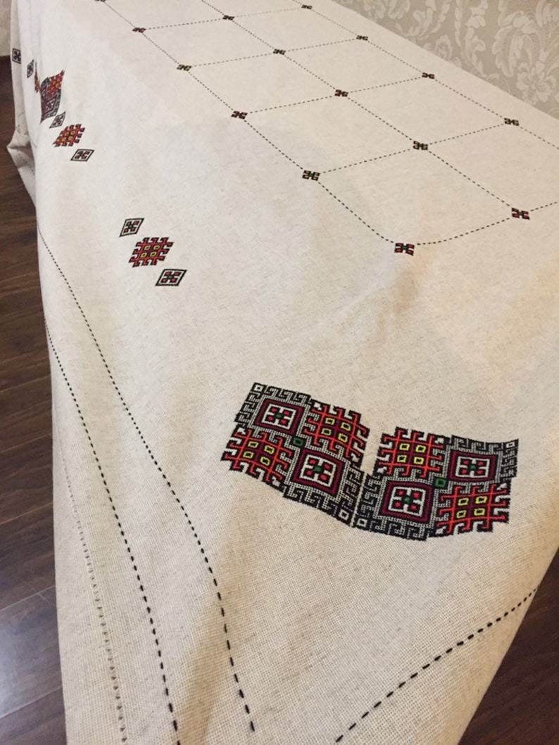 May include: A white linen tablecloth with black and red embroidered geometric patterns. The tablecloth has a black stitched border.