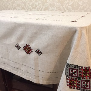 May include: A white linen tablecloth with red, black, and yellow embroidered geometric patterns. The tablecloth has a simple, elegant design and would be perfect for a formal dinner party.