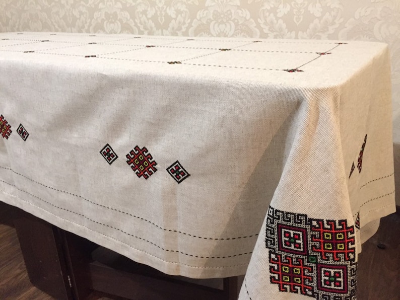 May include: A white linen tablecloth with red, black, and yellow embroidered geometric patterns. The tablecloth has a simple, elegant design and would be perfect for a formal dinner party.