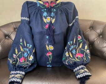 Op de afbeelding: Een marineblauwe linnen blouse met geborduurde bloemenmotieven. De borduursels bevatten verschillende bloemen, waaronder madeliefjes, korenbloemen en roze bloesems. De blouse heeft lange mouwen met een geplooide manchet en een kwastje aan de halslijn.