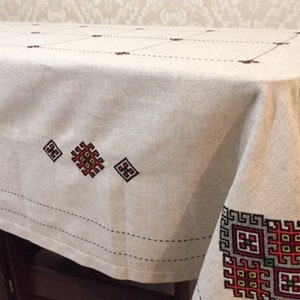 May include: A white linen tablecloth with embroidered red, black, and yellow geometric patterns. The tablecloth has a simple, elegant design and would be perfect for a formal dinner party.