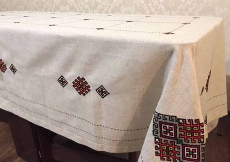 May include: A white linen tablecloth with embroidered red, black, and yellow geometric patterns. The tablecloth has a simple, elegant design and would be perfect for a formal dinner party.