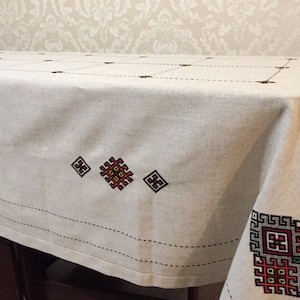 May include: A white linen tablecloth with embroidered red, green, yellow, and black geometric patterns. The tablecloth has a subtle grid pattern and a black border.