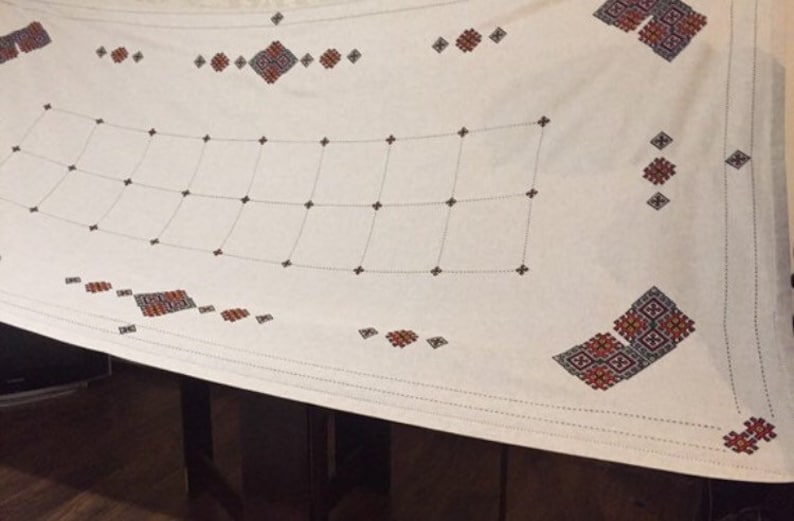 May include: White tablecloth with a grid pattern and embroidered red, black, and white geometric designs.