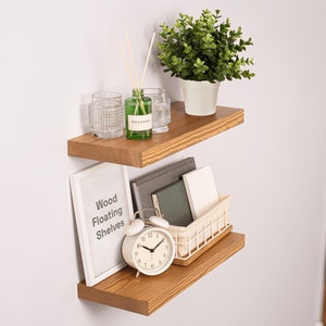 May include: Two wooden floating shelves with a white alarm clock, a small white wire basket, books, a green glass bottle with a label that says "Tea & Ginger", and two clear glass candle holders on the top shelf. The shelves are mounted on a white wall. The top shelf has a small green potted plant with green leaves.