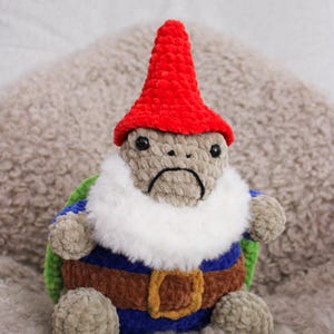May include: A handmade plush gnome turtle with a red hat, white beard, and a sad expression. The turtle wears a blue, brown, and green outfit with a gold buckle. The plush is made of soft, textured yarn.