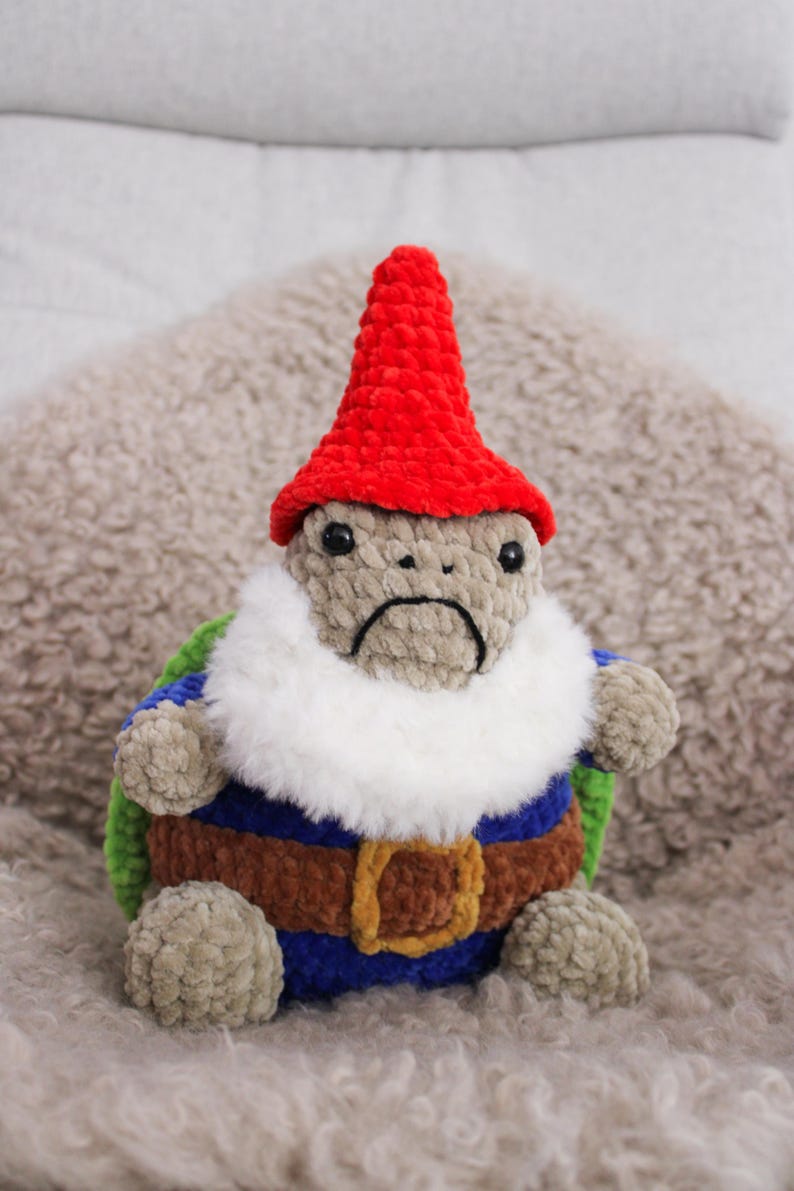 May include: A handmade plush gnome turtle with a red hat, white beard, and a sad expression. The turtle wears a blue, brown, and green outfit with a gold buckle. The plush is made of soft, textured yarn.