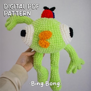 Könnte beinhalten: Ein handgefertigtes grünes Häkelmonster mit großen, weiß umrandeten Augen und orangefarbenen Akzenten. Das Monster hat ein rotes und blaues Kopfstück und der Text "DIGITAL PDF PATTERN" und "Bing Bong" sind sichtbar.