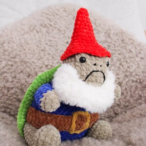 May include: A handmade crochet gnome doll with a red hat, white beard, and a blue and brown outfit. The gnome has a sad expression and is sitting on a soft, textured surface.