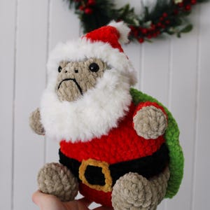 May include: A handmade plush toy of a monkey dressed as Santa Claus. The monkey wears a red suit with a black belt and a white fluffy beard and hat. The toy is beige, red, black, and green. A Christmas wreath is in the background.