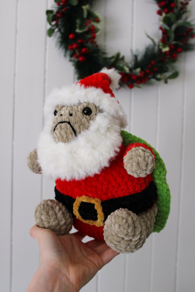 May include: A handmade plush toy of a monkey dressed as Santa Claus. The monkey wears a red suit with a black belt and a white fluffy beard and hat. The toy is beige, red, black, and green. A Christmas wreath is in the background.