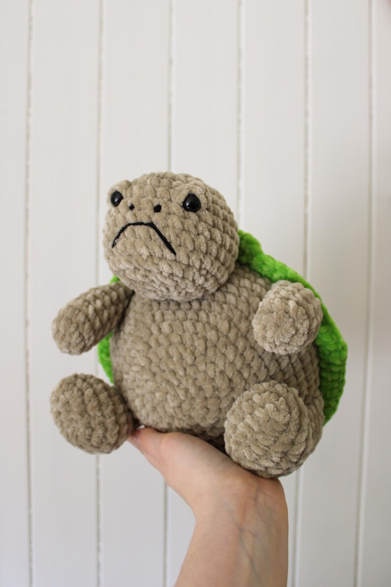 May include: A handmade plush turtle in shades of tan and green. The turtle has a sad expression with black embroidered eyes and a downturned mouth. The shell is green, and the body is a soft, textured tan material.