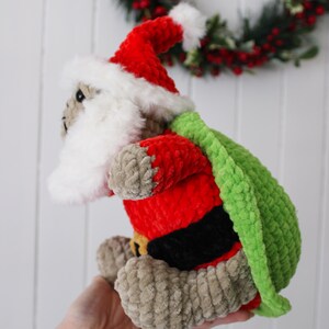 May include: A handmade, plush Santa Claus doll with a red suit, white beard, and green sack. The doll has a red hat with white trim and a black belt. The doll is made of soft, textured yarn and is a festive holiday decoration.