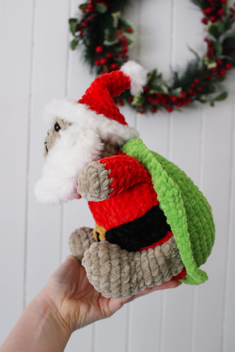 May include: A handmade, plush Santa Claus doll with a red suit, white beard, and green sack. The doll has a red hat with white trim and a black belt. The doll is made of soft, textured yarn and is a festive holiday decoration.