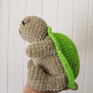 May include: A handmade plush turtle in a seated position. The turtle has a light brown body and a bright green shell. The plush toy is made of a soft, textured material. The turtle's eyes are black.