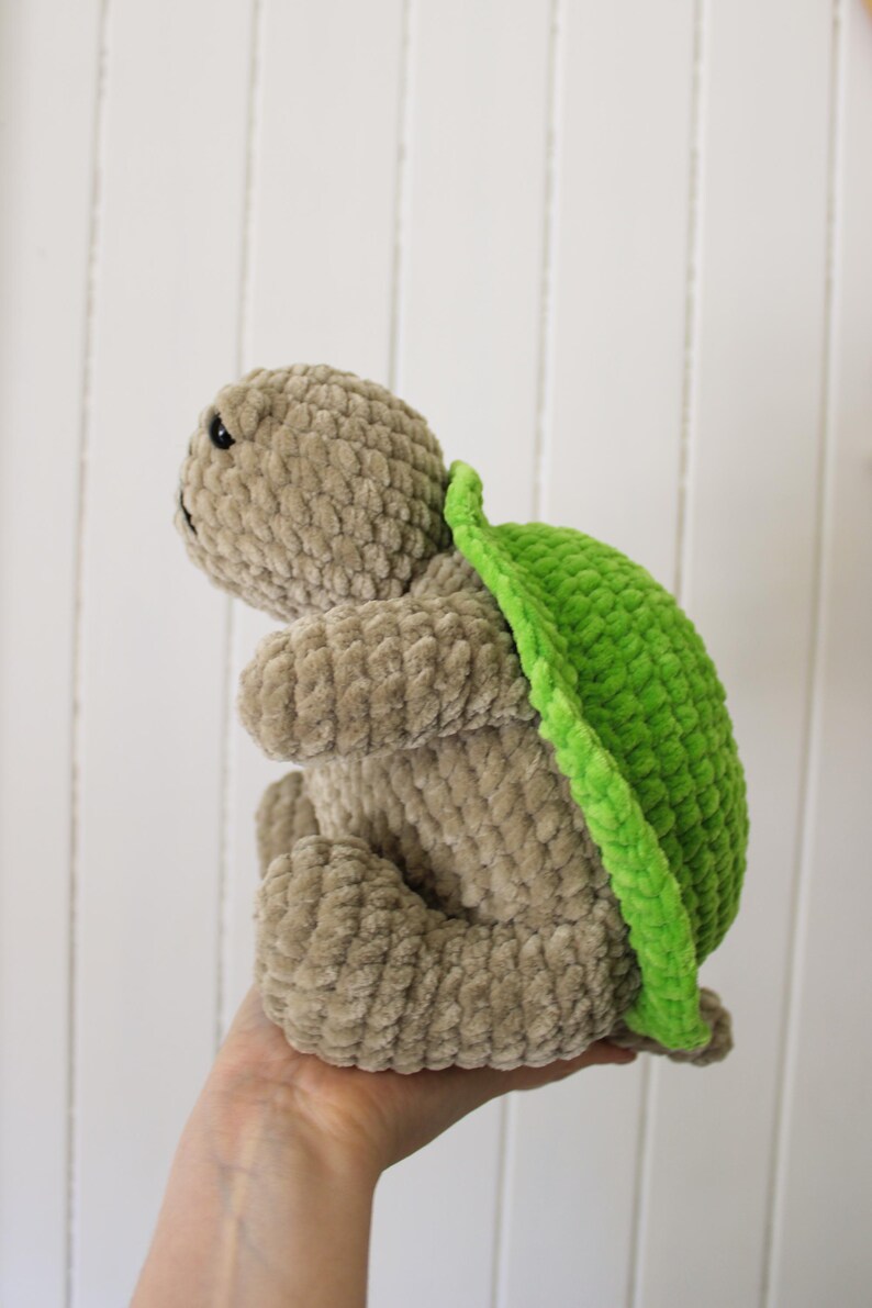 May include: A handmade plush turtle in a seated position. The turtle has a light brown body and a bright green shell. The plush toy is made of a soft, textured material. The turtle's eyes are black.