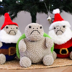 May include: Three crocheted turtle plushies, each with a sad expression. One is dressed as a gnome in a red hat and blue outfit, another is plain gray, and the third is in a Santa Claus outfit with a red hat and white beard.