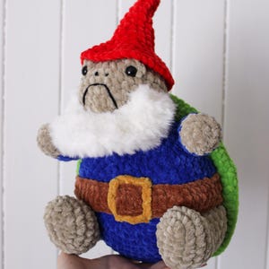 May include: A handmade plush gnome with a red hat, white beard, and blue tunic. The gnome has a brown belt with a gold buckle and green accents. The gnome is approximately 8 inches tall.