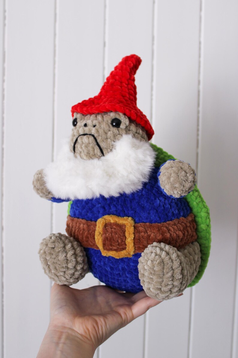 May include: A handmade plush gnome with a red hat, white beard, and blue tunic. The gnome has a brown belt with a gold buckle and green accents. The gnome is approximately 8 inches tall.
