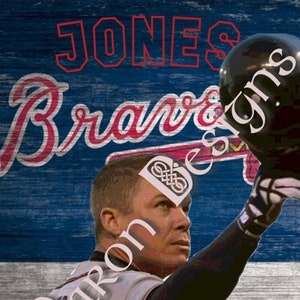 May include: A baseball player wearing a grey jersey with the number 10 on the front, a red and blue baseball cap, and a black glove. The player is standing in front of a red, white, and blue American flag with the words "Braves" and "JONES" in red letters.