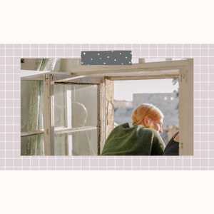 May include: A person with red hair sits by a window looking out at a cityscape. The window is old and has multiple panes. The person is wearing a green sweater.