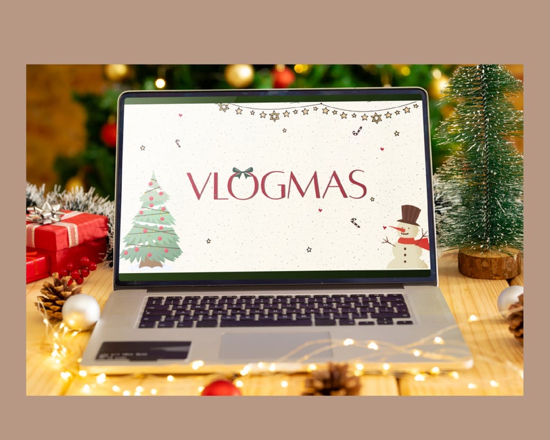 May include: A laptop computer with a white screen displaying the text "VLOGMAS" in red letters. The screen also shows a cartoon Christmas tree and a snowman. The laptop is on a wooden table with a small artificial Christmas tree and string lights.