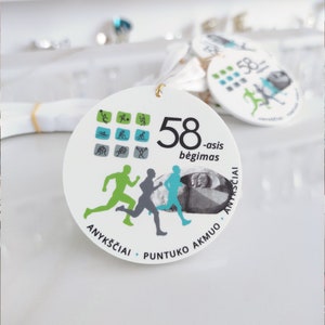 Può includere: Un'etichetta circolare bianca con il numero "58" e le parole "-asis bėgimas" stampate in nero. L'etichetta presenta figure di corridori in verde, grigio e blu, insieme a piccole icone sportive. È presente anche il testo "ANYKŠČIAI • PUNTUKO AKMUO".
