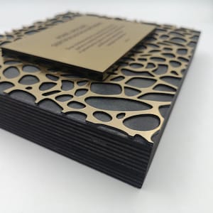 Puede incluir: Una pieza decorativa cuadrada y en capas con una base negra y una superposici&oacute;n de patr&oacute;n org&aacute;nico dorado. Una pieza rectangular m&aacute;s peque&ntilde;a de color dorado descansa encima, con texto visible. La pieza tiene una est&eacute;tica moderna y art&iacute;stica.