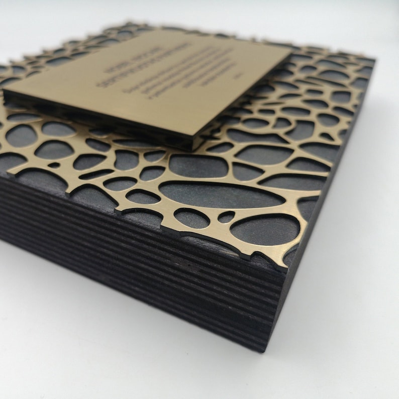 Puede incluir: Una pieza decorativa cuadrada y en capas con una base negra y una superposici&oacute;n de patr&oacute;n org&aacute;nico dorado. Una pieza rectangular m&aacute;s peque&ntilde;a de color dorado descansa encima, con texto visible. La pieza tiene una est&eacute;tica moderna y art&iacute;stica.