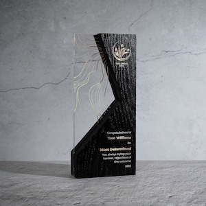 Puede incluir: Un premio rectangular con una parte delantera transparente y una parte trasera de madera negra. El premio tiene detalles dorados y el texto "Most Determined" y "Wellness Festival". El premio es para Tom Williams.