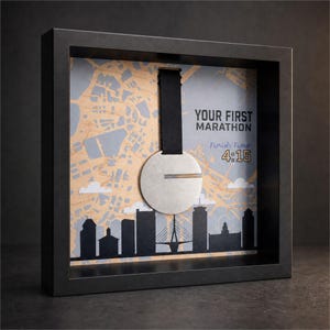 Op de afbeelding: Een zwarte schaduwdooslijst met een zilveren ronde medaille aan een zwart lint. De achtergrond toont een beige en grijze kaart met een zwart silhouet van een stad, de tekst YOUR FIRST MARATHON en Finish Time 4:15.