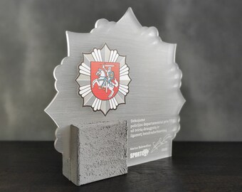 Könnte beinhalten: Eine transparente Acryl-Auszeichnung mit einem grauen Betonfuß. Die Auszeichnung zeigt ein Wappenemblem und Text in einer Fremdsprache. Die Auszeichnung ist etwa 15 cm hoch.