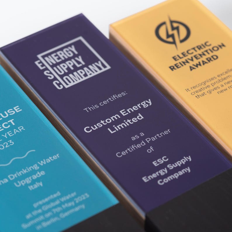 May include: Close-up of three rectangular awards with black bases. The awards are blue, purple, and gold. The purple award has the text "ENERGY SUPPLY COMPANY" and "Custom Energy Limited". The gold award reads "ELECTRIC REINVENTION AWARD".