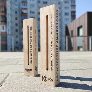 Könnte beinhalten: Zwei Holzpreise mit eingraviertem Text. Einer lautet „MITARBEITER DES MONATS“ und „AUSZEICHNUNG 2023“. Der andere lautet „ARCHITEKT DES JAHRES“ und „ARCHITEKTURPREISE 2023“. Beide haben ein Logo und sind hellbraun.