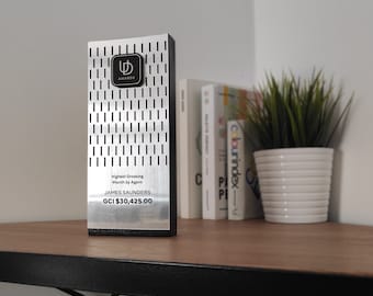 May include: A silver award plaque with a black logo and the text "ゆ AWARDS". The plaque has a black base and is engraved with the text "Highest Grossing Month by Agent", "JAMES SAUNDERS", and "GCI $30,425.00".