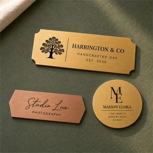 Könnte beinhalten: Drei individuelle Metallschilder mit graviertem Text und Logos. Ein rechteckiges goldenes Schild trägt die Aufschrift Harrington & Co Handcrafted Oak Est. 2026, ein kupferfarbenes sechseckiges Schild Studio Lux Photography und ein rundes goldenes Schild Maison Elora.