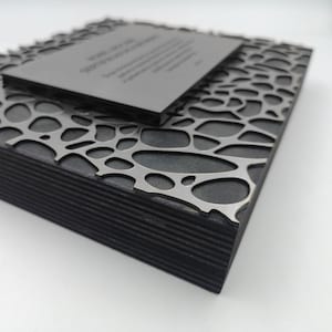 Puede incluir: Una caja de madera gris oscuro con una superposici&oacute;n met&aacute;lica calada decorativa. Una placa rectangular m&aacute;s peque&ntilde;a descansa encima, con texto grabado. La caja tiene una construcci&oacute;n en capas y el dise&ntilde;o es moderno y geom&eacute;trico.