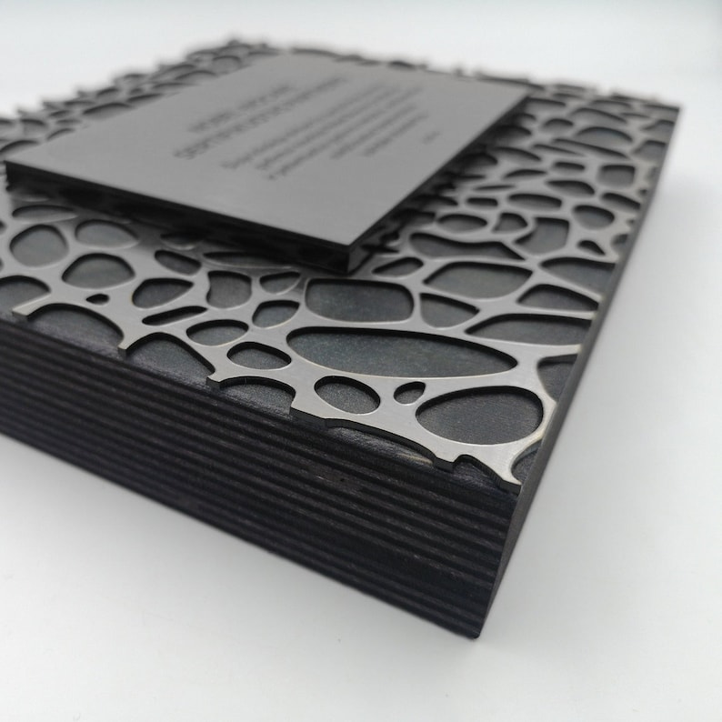 Puede incluir: Una caja de madera gris oscuro con una superposici&oacute;n met&aacute;lica calada decorativa. Una placa rectangular m&aacute;s peque&ntilde;a descansa encima, con texto grabado. La caja tiene una construcci&oacute;n en capas y el dise&ntilde;o es moderno y geom&eacute;trico.