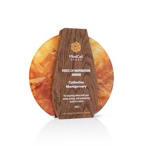 Może przedstawiać: Okrągła tablica pamiątkowa z ciemnobrązowym drewnianym panelem środkowym z napisem MindCell GROUP, VOICE OF INSPIRATION AWARD, Catherine Montgomery i 2025, otoczona dwoma półprzezroczystymi, bursztynowymi dyskami z żywicy o teksturowanej powierzchni.