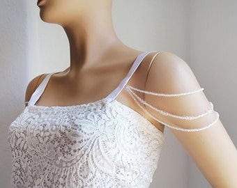 May include: A white lace top featuring thin shoulder straps and a decorative multi-strand beaded arm chain. The arm chain consists of three draped layers of small, clear, reflective beads that connect to the shoulder strap of the garment.