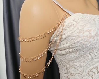 May include: Gold-toned shoulder jewelry with three cascading strands. Each strand is adorned with small, dangling embellishments. The jewelry is draped over a mannequin wearing a white lace dress, suggesting bridal or formal wear.