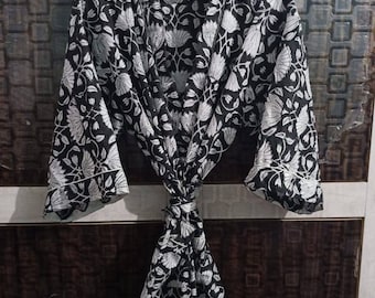 May include: A black and white floral print robe hanging on a wooden hanger. The robe features a repeating pattern of stylized flowers and leaves. The robe is tied at the waist with a matching fabric belt. The sleeves have white trim.
