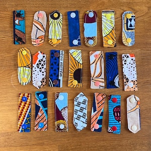 Könnte beinhalten: Eine Sammlung von 18 bunten Stoff-Maskenverlängerungen mit verschiedenen Mustern, darunter geometrische, florale und abstrakte Designs. Jeder Verlängerer hat zwei Knöpfe zum Befestigen an einer Maske.